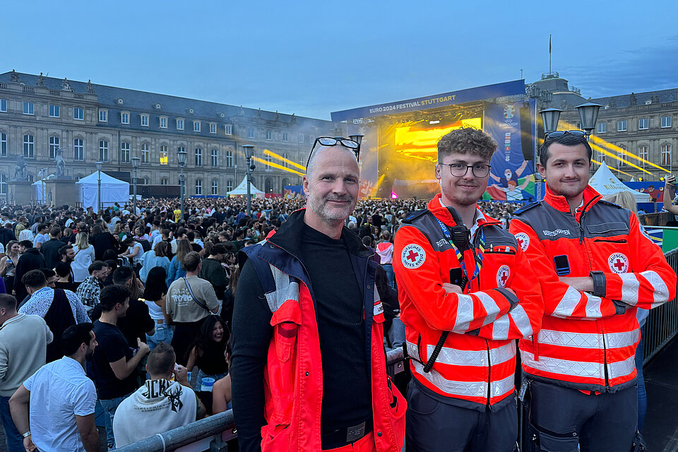 Drei Mitglieder des Deutschen Roten Kreuzes stehen beim EURO 2024 Festival in Stuttgart vor der Menschenmenge. Sie tragen rote Rettungsuniformen und lächeln in die Kamera.