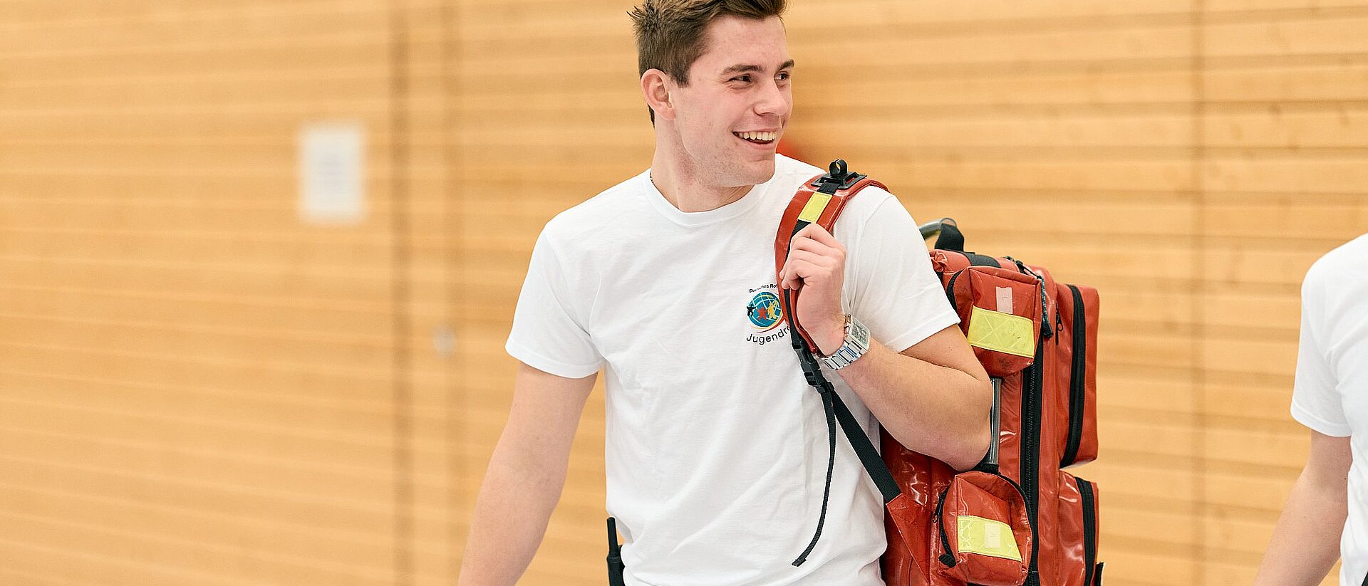 Ein junger Mann mit JRK-T-Shirt hält einen Erste-Hilfe-Rucksack über der Schulter und lächelt