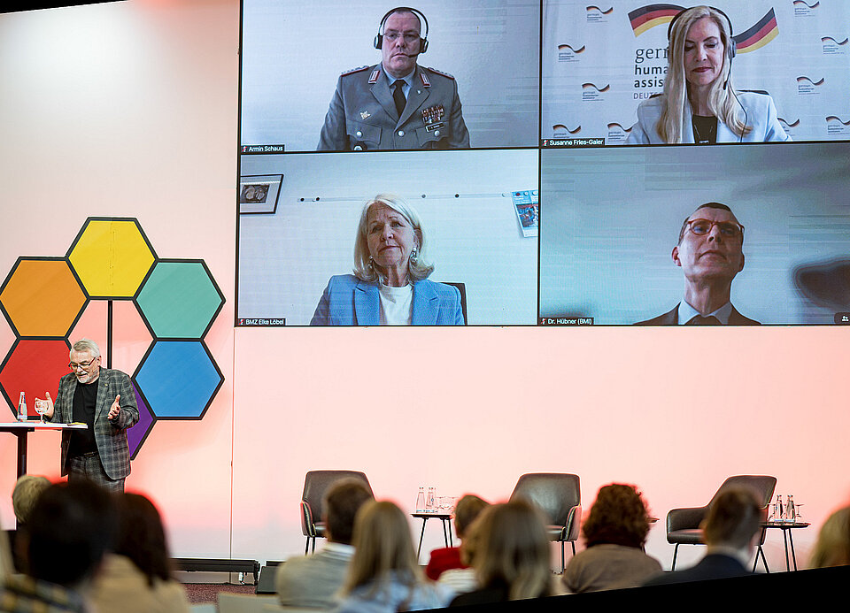Ein Sprecher steht auf einem Podium, im Sall sitzen Zuschauer und auf dem Bildschirm sind vier virtuelle Teilnehmer projeziert.