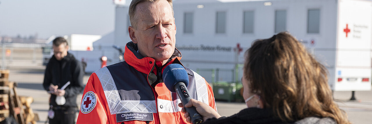 DRK-Generalsekretär Christian Reuter steht in DRK-Einsatzkleidung im Freien und wird von einer Frau  interviewt