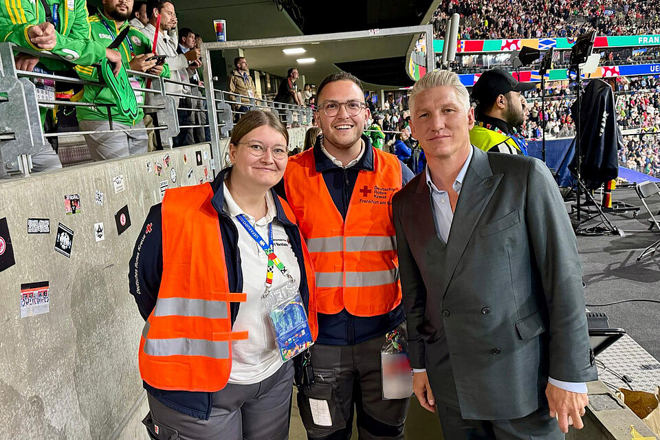 Zwei Mitglieder des Deutschen Roten Kreuzes in orangefarbenen Westen mit Ex-Fußballprofi Bastian Schweinsteiger in einem vollbesetzten Stadion.