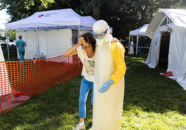 Eine Frau wird von einem Mann in Schutzkleidung gestützt. Im Hintergrund Zelte des Feldkrankenhauses