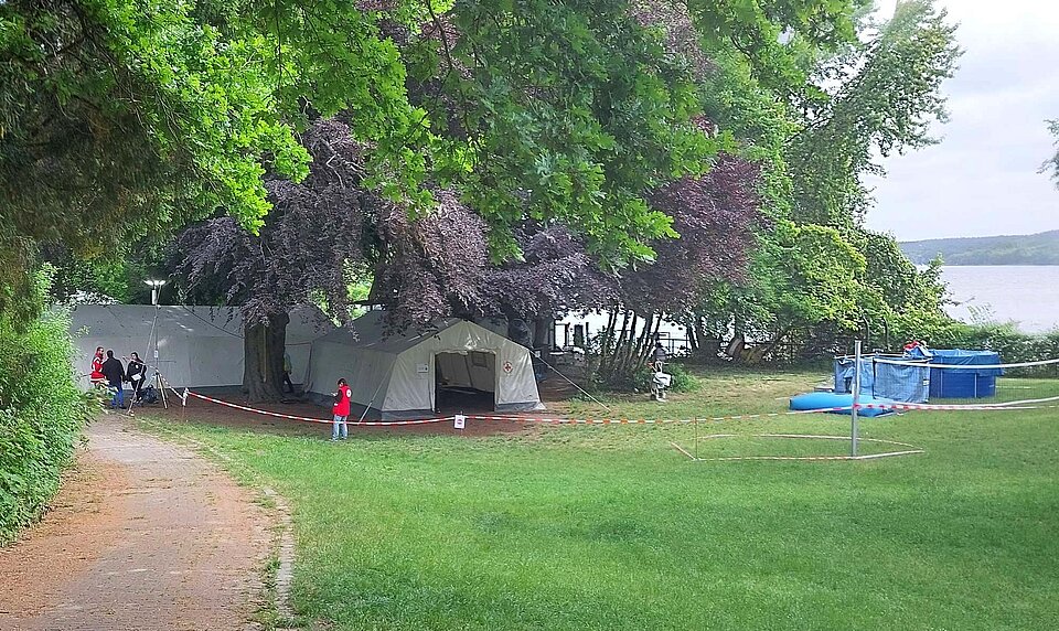 DRK-Surge-Uebung_auf_Schwanenwerder Auf einer Wiese am Wasser unter Bäumen stehen Zelte und eine mobile Trinkwasseranlage. Menschen mit und ohne Rotkreuzwesten stehen auf der Wiese