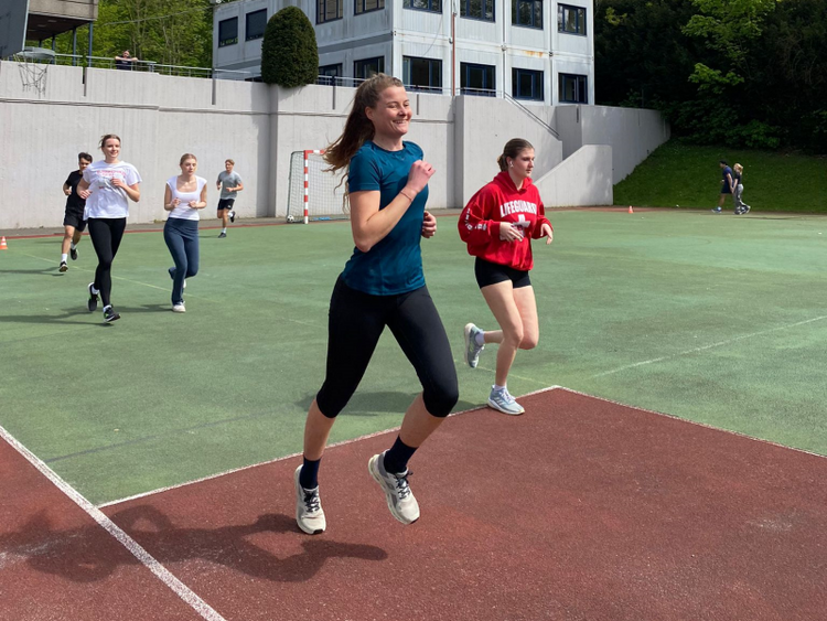 Spendenlauf an der Internationalen Deutschen Schule in Brüssel Das Bild zeigt einige Schülerinnen und Schüler, die anlässlich des Spendenlaufs der internationalen deutschen Schule in Brüssel um einen Sportplatz im Freien rennen. Die Sonne scheint, im Hintergrund sieht man hinter einer Mauer Schulgebäude. Die junge Läuferin im Vordergrund lächelt beim Laufen.