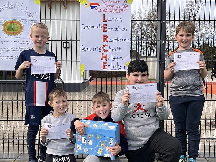 Spendenlauf der Grundschule am Lerchenweg Auf dem Bild sind vier Schüler der Grundschule am Lerchenweg aus Monheim (NRW) zu sehen. Sie halten ihre ausgefüllten Spendenlauf-Zettel in den Händen und lächeln stolz in die Kamera. Sie tragen Sportsachen. Im Hintergrund hängt das Motto der Grundschule: Lernen, füreinander einstehen, respektieren, Gemeinschaft, helfen, erleben.
