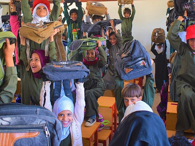 Man sieht viele Schulkinder in einem Raum, die lachend ihre Schulranzen in die Höhe halten.