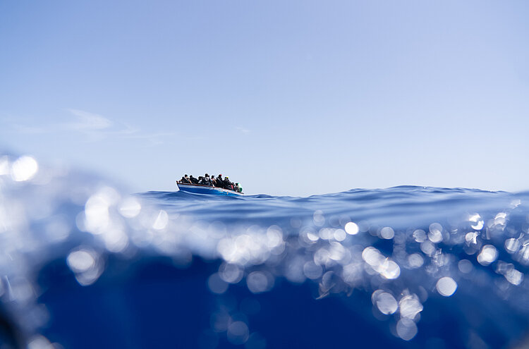 Seenotrettung_im_Mittelmeer.jpg Man sieht im Vordergrund die Meereswellen, im Hintergrund ein kleines Boot.