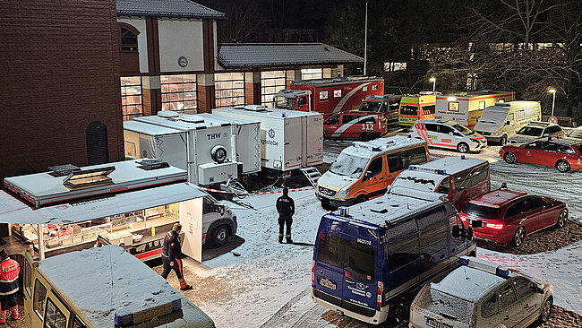 Nachtszene vor einer Feuerwache mit Schnee auf dem Boden und auf den Fahrzeugen. Mehrere Einsatzfahrzeuge von Feuerwehr, Rettungsdienst, Polizei und THW stehen dicht beieinander während Einsatzkräfte zwischen ihnen gehen und ein beleuchteter Verpflegungswagen auf einen laufenden Großeinsatz hinweist.