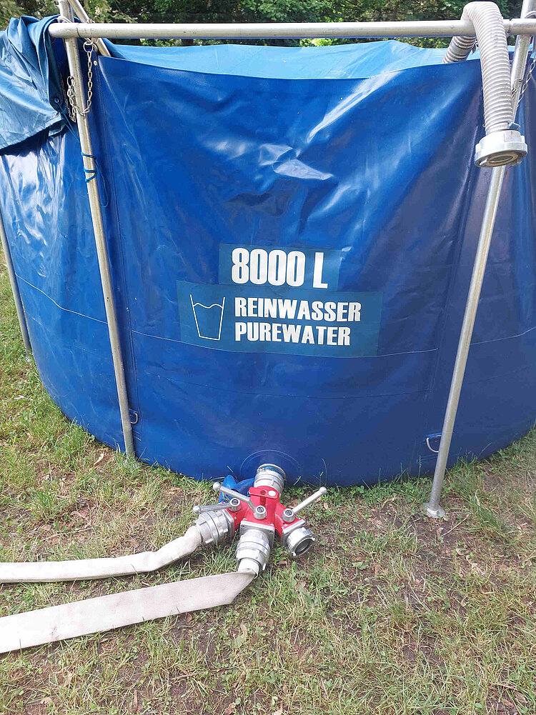 Trinkwasseraufbereitung_des_DRK Aus einem Kunststofftank mit der Aufschrift "(8000 Liter Reinwasser Pure Water" ragen mehrere dicke Schläuche