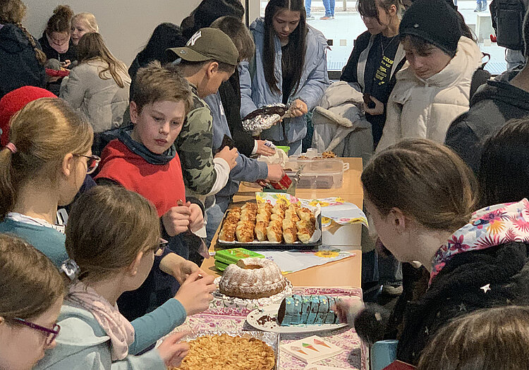 Spendenaktion_Copyright_Ernst_Mach_Gymnasium_Huerth Das Bild zeigt viele Schülerinnen und Schüler, die um einen Tisch herumstehen. Auf der einen Seite stehen Kinder aus einer fünften Klasse, die Kuchen verkaufen. Auf der anderen Seite stehen Schüler unterschiedlicher Klassenstufen, um sich Kuchen zu kaufen.