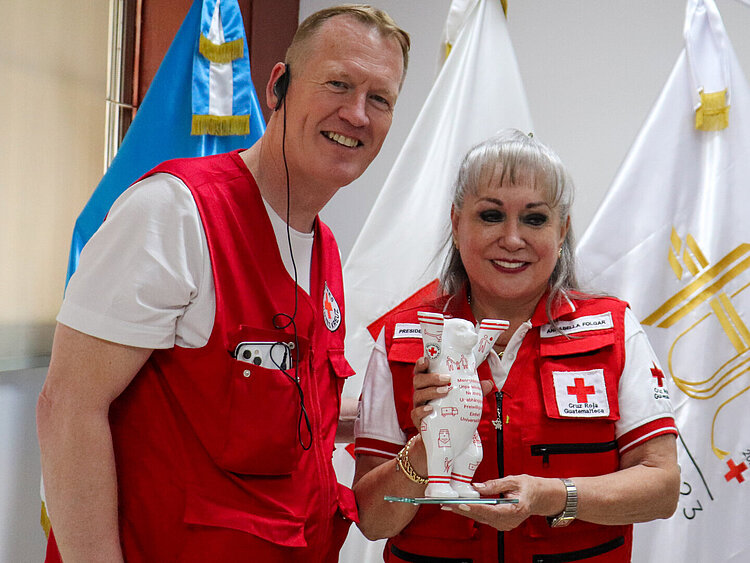 Christian Reuter und die Präsidentin des Guatemaltekischen Roten Kreuzes, Annabella Folgar Bonilla. Christian Reuter und Annabella Folgar Bonilla, in Rote-Kreuz-Westen gekleidet, stehen lächelnd vor Flaggen. Die Frau hält einen Buddy Bear aus Porzellan mit Rote-Kreuz-Emblemen..