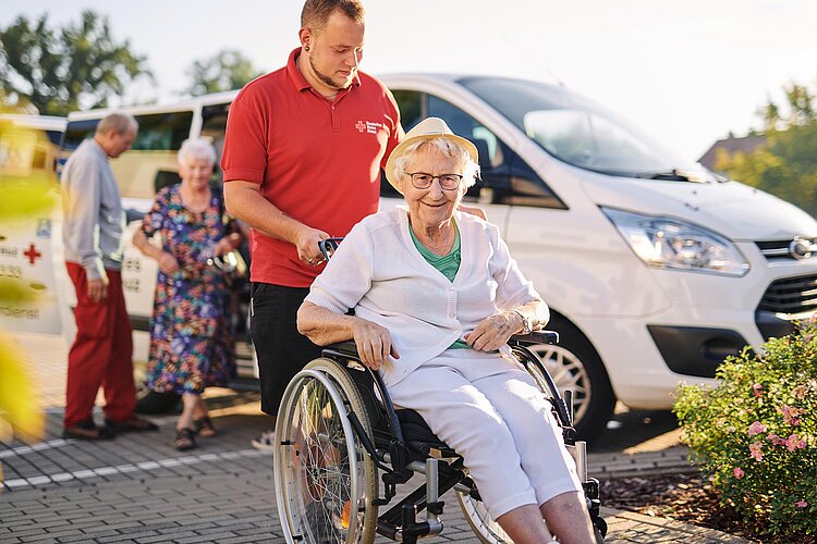 Tagespflege: Ankunft mit Fahrdienst DRK-Pfleger und Seniorin im Rollstuhl vor DRK-Bus