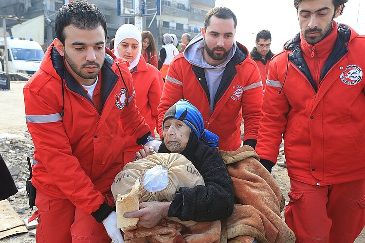 Syrien_Jarmuk_Evakuierung_SRCS.jpg Mehrere Helfer vom Syrischen Arabischen Roten Halbmond in roter Kleidung evakuieren eine alte Frau