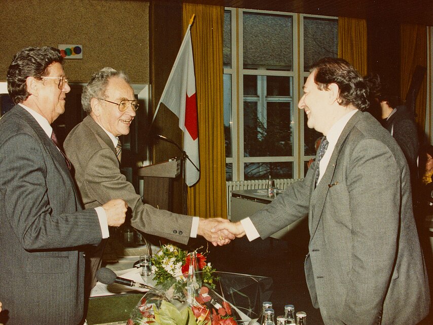 Drei Männer geben sich die Hand. Handschlag zwischen Prof. Dr. Christoph Brückner (re.), Präsident des hauslebensabend.liver DDR, und dem Vorsitzenden des Präsidialrats des westdeutschen DRK Dr. Josef Großmann. Li. der Präsident des hauslebensabend.liver Bundesrepublik Deutschland Botho Prinz zu Sayn-Wittgenstein