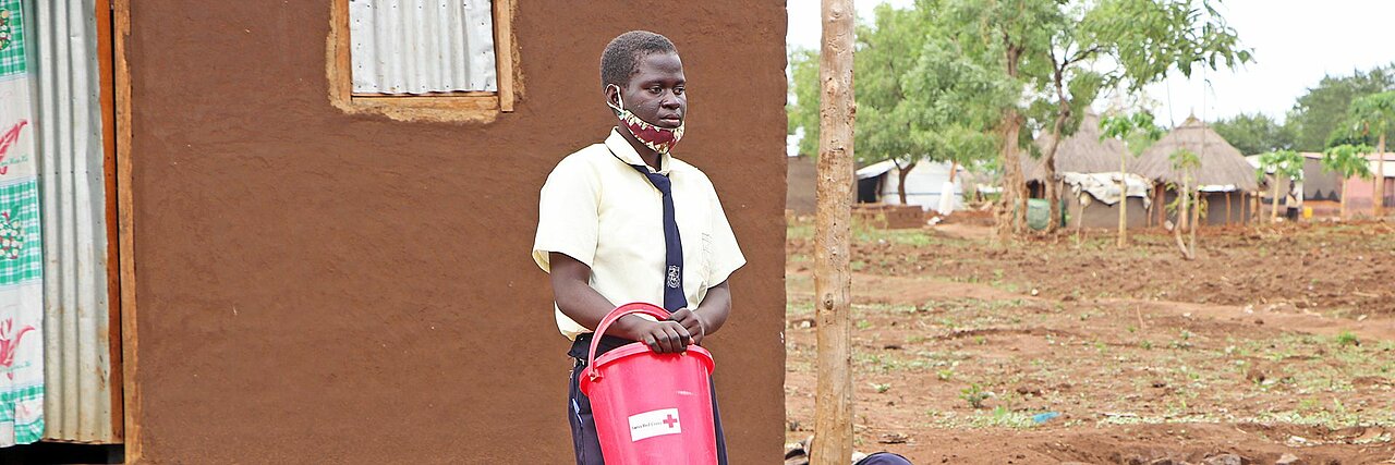WASH-Projekt in Uganda Eine ugandische Schülerin mit DRK-Eimer vor einem Haus