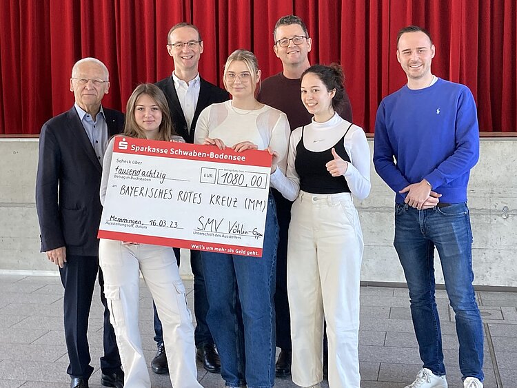 Spendenaktion © Vöhlin-Gymnasium Memmingen Auf dem Bild sind sieben Personen zu sehen. Bei ihnen handelt es sich um (von links nach rechts) hintere Reihe: Dr. Ivo Holzinger, Vorstand vom DRK Kreisverband Unterallgäu, Heinz Scheitzender vom KV Unterallgäu, Christian Herrmann (Schulleiter Vöhlin-Gymnasium) und Konstantin Heiß (Lehrer Vöhlin-Gymnasium). Alle vier Männer sind weiß und lächeln in die Kamera, drei von ihnen tragen Brillen, alle haben kurze Haare. Im Vordergrund stehen von links nach rechts die drei Schülersprecherinnen des Gymnasiums. Sie halten den sehr großen Scheck über 108 Euro in der Hand und lächeln in die Kamera.