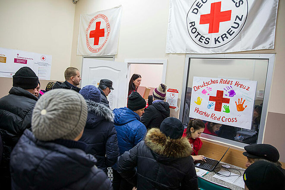 Das Ukrainische Rote Kreuz verteilt Bargeldhilfen unterstützt vom DRK Viele Menschen stehen vor einem Tisch. Eine Mitarbeiterin des Ukrainischen Roten Kreuzes arbeitet dort am Laptop. An der Wand hängen Banner des Ukrainischen Roten Kreuzes und des DRK.