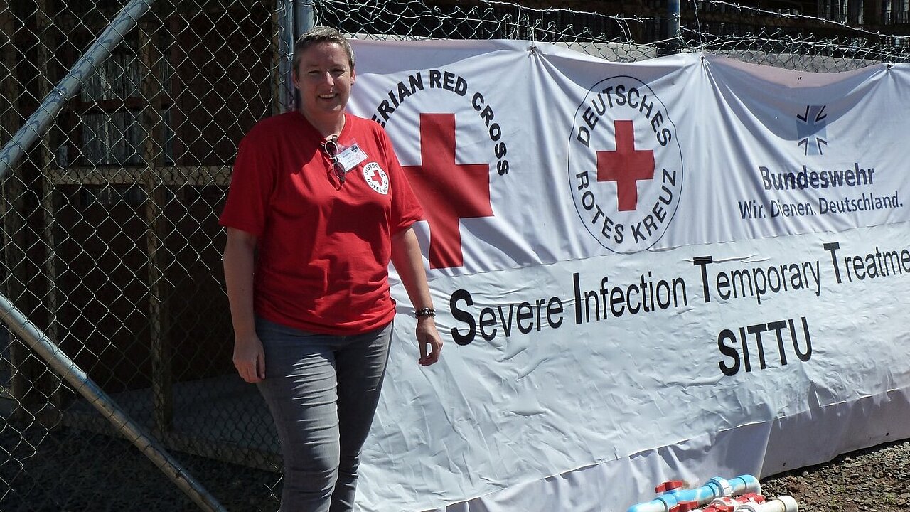 Regine Reim vor dem Ebola-Behandlungszentrum in Monrovia / Liberia Eine Rotkreuz-Mitarbeiterin steht vor einem Zelt, auf dem die Logos des Liberianischen, des Deutrschen Roten Kreuzes und der Bundeswehr zu sehen sind.