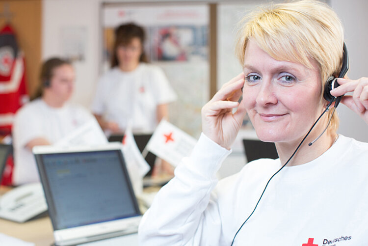 Die Pflegeberatung des DRK. Eine DRK-Mitarbeiterin sitzt vor einem Laptop und trägt ein Headset. Im Hintergrund sind zwei weitere DRK-Mitarbeiterinnen