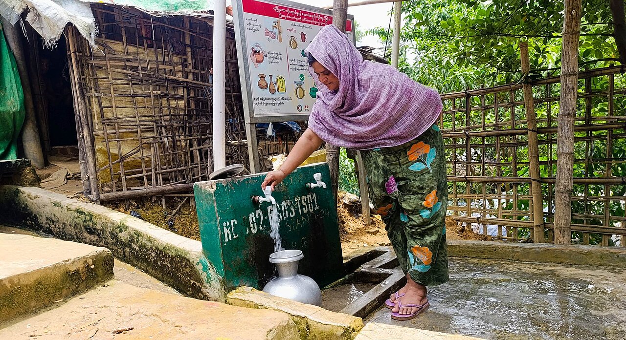 Eine Frau steht draußen an einer Wasserentnahmestelle und lässt Wasser in einen Blechkrug laufen